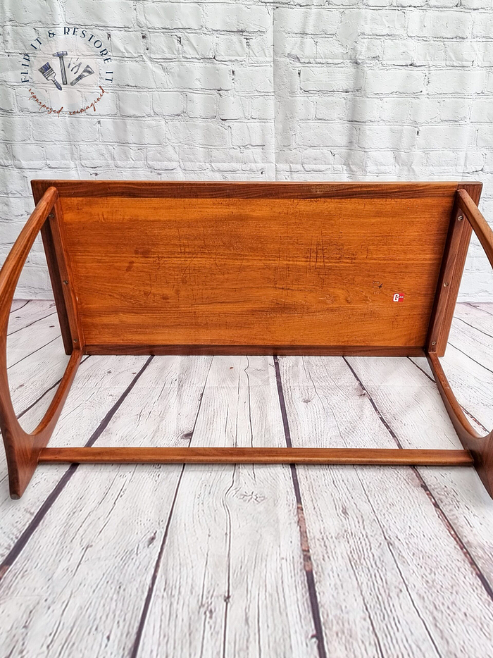 The G Plan Astro Mid Century Modern Vintage Teak Coffee Table from the 1960s is shown upside down on a light-colored wooden floor. The underside of the table's rectangular top is visible, revealing its construction. The table legs extend upward, highlighting their curved design. A decorative logo can be seen on the wall in the background.