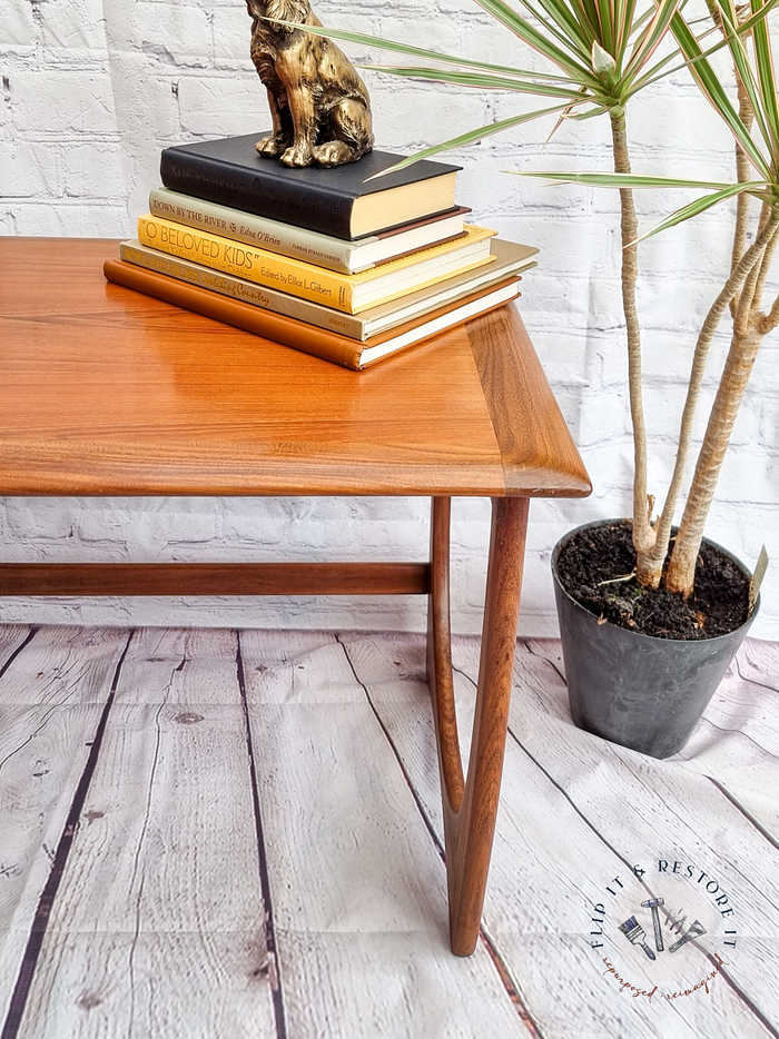 A G Plan Astro Mid Century Modern Vintage Teak Coffee Table from the 1960s holds a stack of books topped with a small gold cat sculpture, sitting next to a potted plant with long, slender leaves. The background features a white brick wall and the floor is made of wooden planks. A logo in the bottom right corner reads "BUILD IT RESTORE IT.