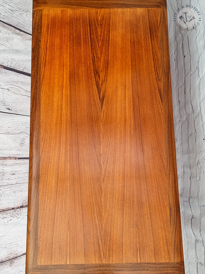 A G Plan Astro Mid Century Modern Vintage Teak Coffee Table from the 1960s, showcasing a smooth, polished surface with a rich, warm brown hue and distinct wood grain patterns. The table is set against a white-washed wooden background that features visible textures and a decorative emblem in the top right corner.