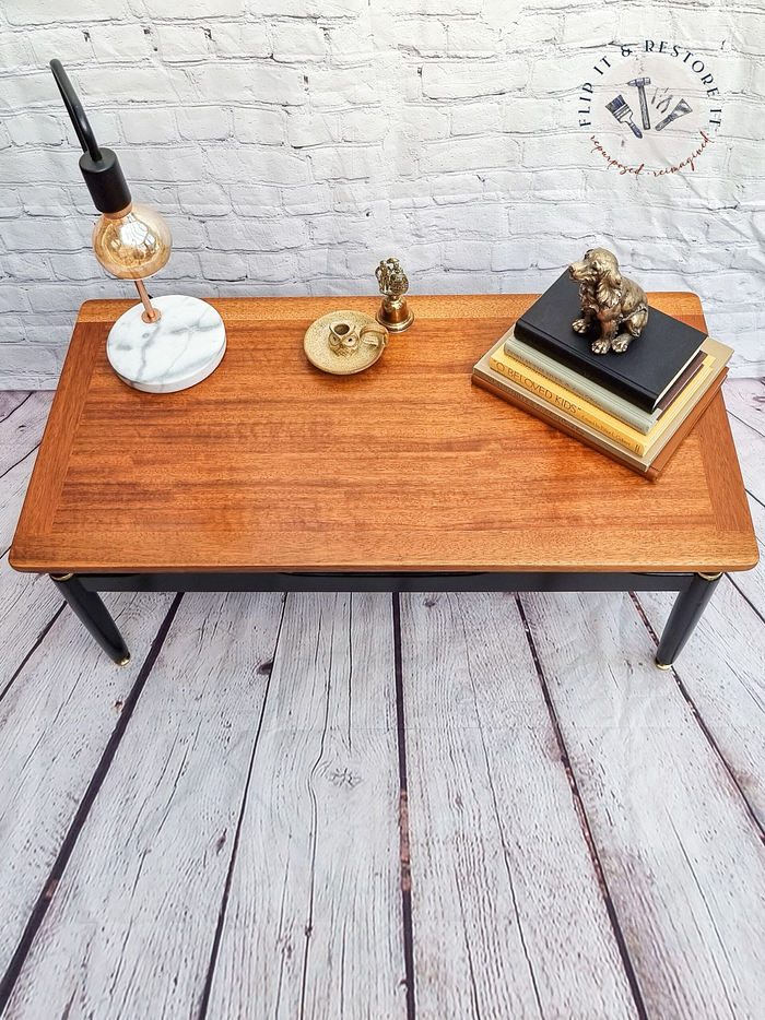 The G-Plan Librenza Mid Century Modern Black and Tola Vintage Coffee Table from the 1950s is adorned with a small, gold-colored tray holding a candle and decor, a vintage lamp with a marble base, and a stack of books topped with a bronze lion statue. The setup is placed on a wooden floor with a brick wall in the background.