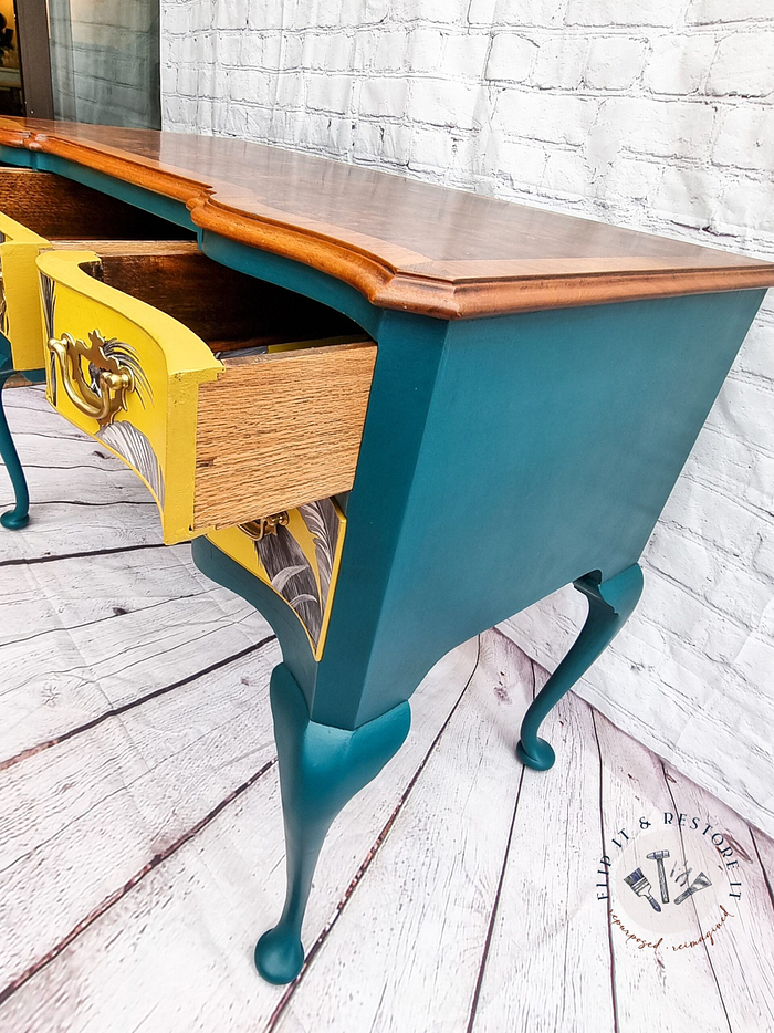 Against a white brick wall, the Queen Anne Painted Burr Walnut Dressing Table/Vanity/Desk/Sideboard showcases its teal-painted legs and sides with a natural wood top. One drawer is partially open, revealing a vibrant yellow interior adorned with decorative patterns and featuring a vintage-style brass handle. An artist's emblem rests on the floor nearby.