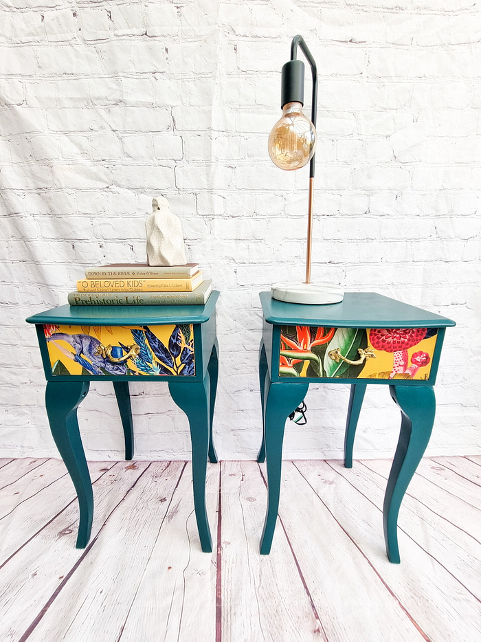 Two pieces from the Queen Anne Painted Burr Walnut Bedroom Set—a dressing table and a matching stool—are positioned against a white brick wall. On the left is the dressing table, adorned with stacked books and a small white animal figurine. To its right, the sleek lamp with an exposed bulb adds a modern touch to the classic setup.