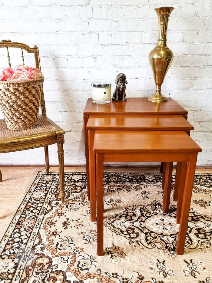 An Ercol Saville Ash Long Coffee Table with Smoked Glass Mid Century Vintage features prominently against a white brick wall, topped with a brass vase, a candle, and a small statuette. To the left of this setup is a wooden chair with a woven seat and a basket of pink roses, all elegantly placed on an ornate patterned rug.