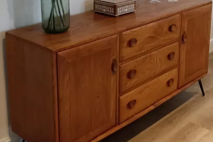 A vintage wooden sideboard with a mid-century modern design. It features two doors and four drawers with round wooden handles. On top of the sideboard are a decorative box, two candleholders, and a tall green vase containing decorative branches.