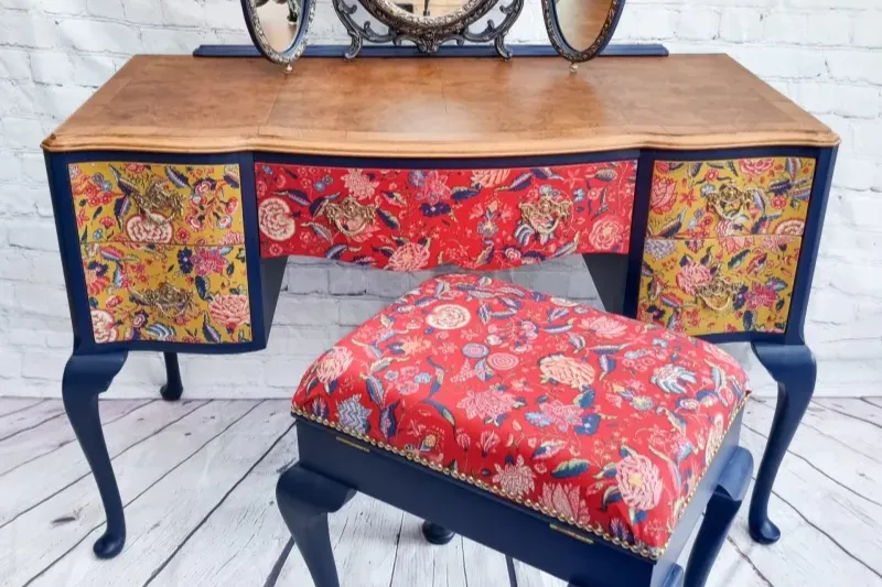 A vintage wooden vanity table with a matching stool. Both feature intricate, colorful floral patterns with predominantly red upholstery. The table has a medium-tone wood surface and dark blue legs, with decorative painted side panels matching the upholstery.