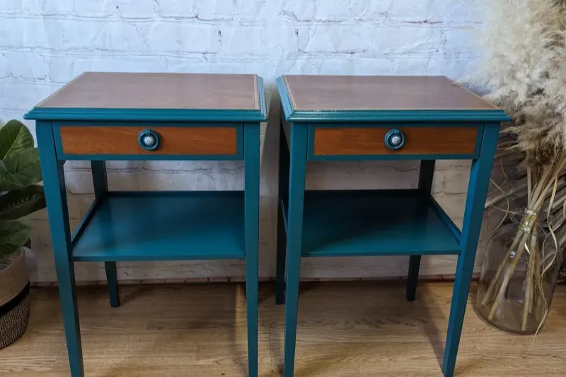 Two matching teal-painted wooden side tables with single drawers featuring brown fronts and round handles. Each table has a lower shelf for storage. They are positioned side by side on a wooden floor, against a white brick wall, with plants and dried pampas grass nearby.