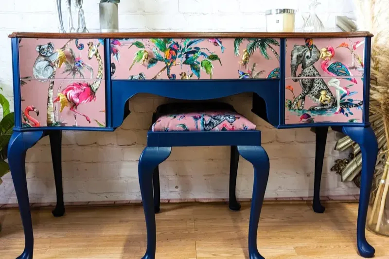 A vintage vanity table with a matching stool, painted in deep blue. The drawers and stool have pink fronts adorned with vibrant illustrations of various birds and animals, including flamingos, parrots, zebras, koalas, and monkeys, on each drawer surface.