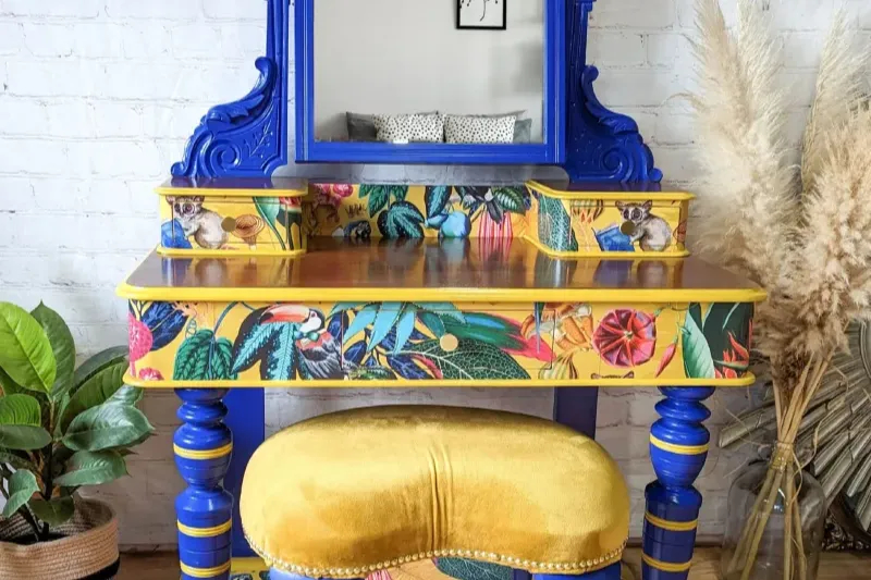 A vibrant blue vanity table with intricately painted tropical designs and animal motifs stands against a white brick wall. A yellow cushioned stool with ornate details matches the vanity. Potted plants and pampas grass decorate the surrounding area.