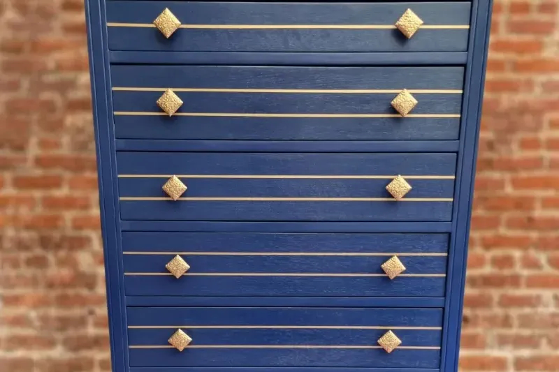 A tall blue dresser with seven drawers, each adorned with gold horizontal handles and square-shaped gold knobs. The dresser stands against a background of red brick, giving a modern and stylish appearance.