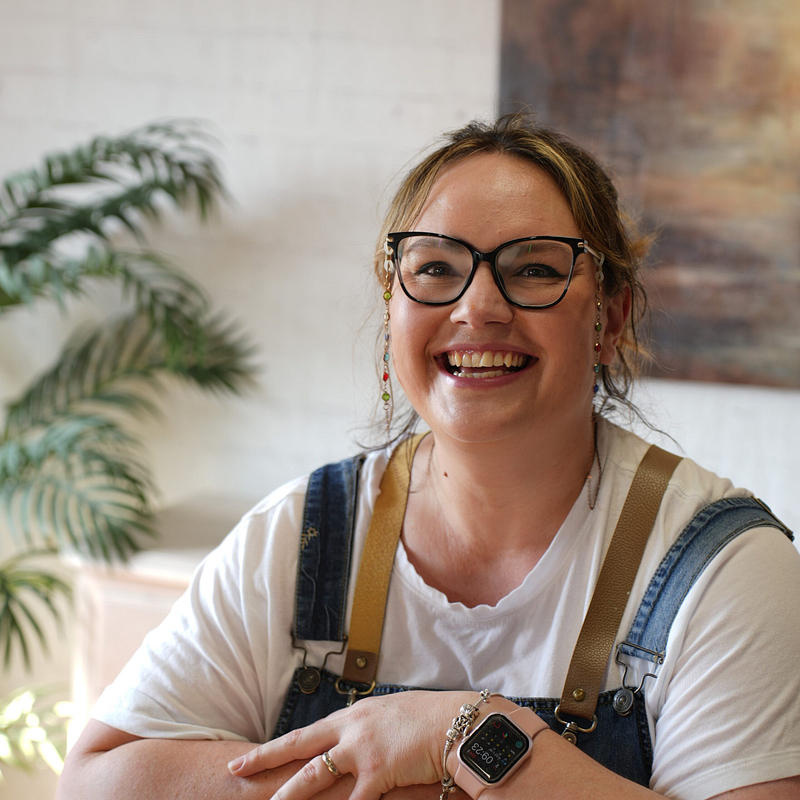 A smiling person wearing glasses and a smartwatch sits at a table. They are dressed in a white shirt and denim overalls. In the background, there is a leafy plant and a blurred painting on the wall. The setting appears to be a warmly lit indoor space.