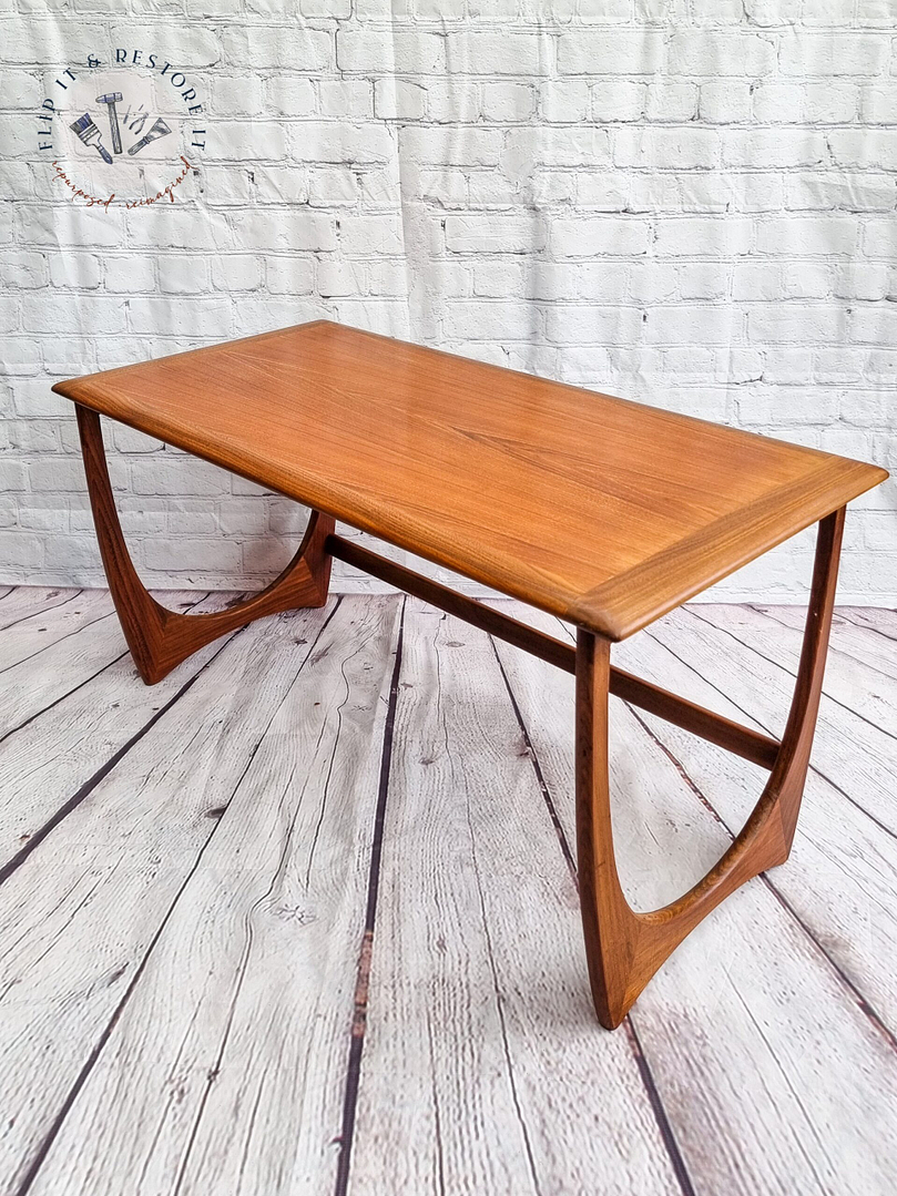 The G Plan Astro Mid Century Modern Vintage Teak Coffee Table from the 1960s showcases a sleek, minimalist design with a rectangular top featuring rounded edges and elegantly curved legs. The table is set against a backdrop of white brick walls and wooden floorboards.