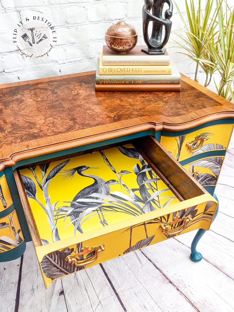 The Queen Anne Painted Burr Walnut Dressing Table/Vanity/Desk/Sideboard is adorned with books and a sculpture. One drawer is open, revealing a vibrant yellow interior with a black and white bird and plant design. The desk features teal accents and brass handles, set against a white brick wall and sumac flooring.